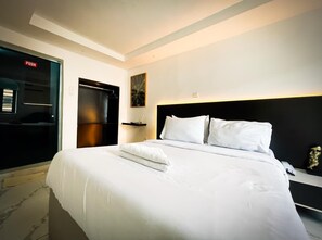 A hotel room with a bed, a bedside table, a nightstand, a wall-mounted shelf, a painting, and a door with a push sign. - LUXE APARTMENTS (Lagos)