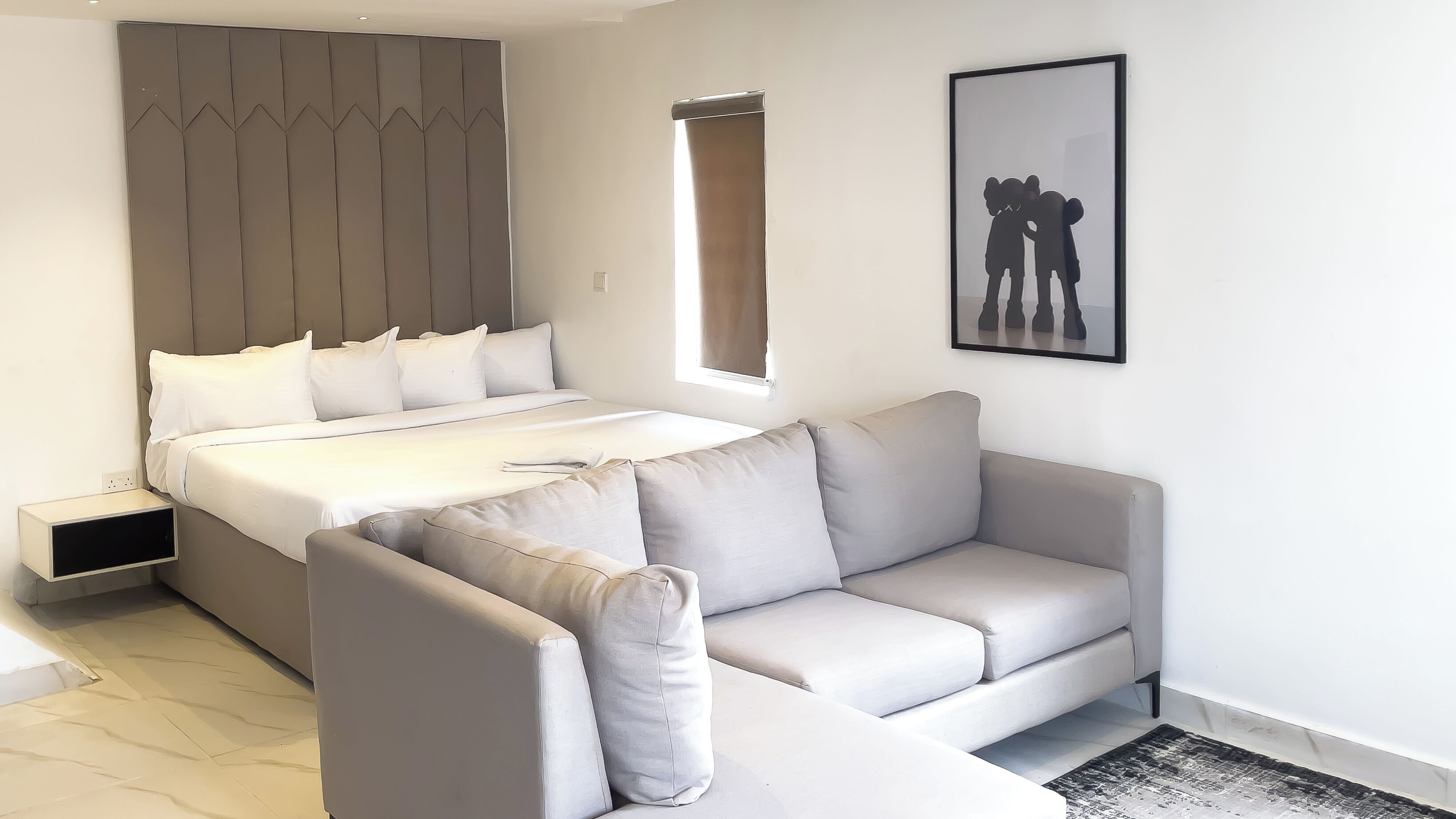 A modern bedroom with a bed, a grey sofa, a small bedside table, and a window.