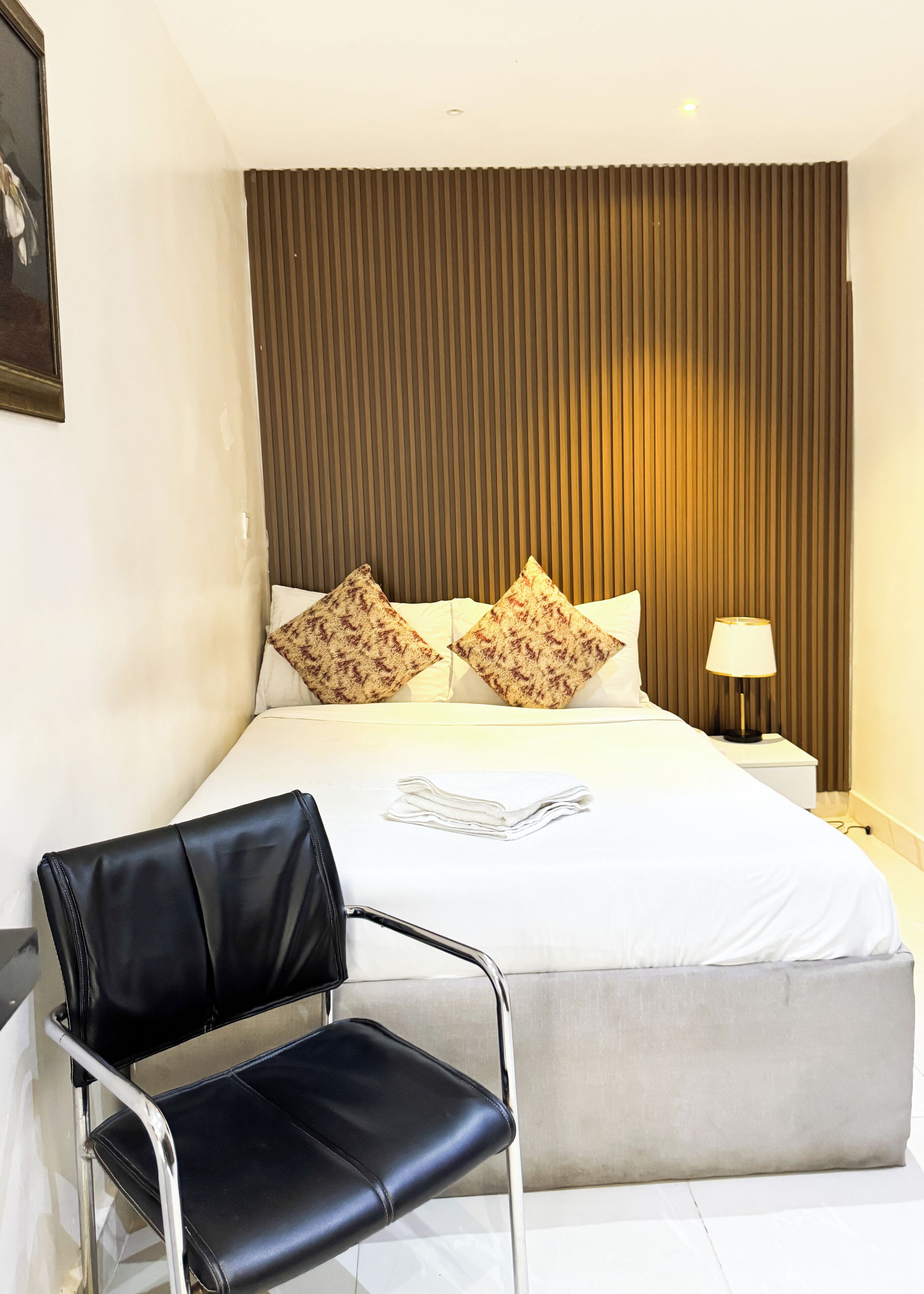 Una camera d'hotel con un letto, cuscini, una sedia, un comodino con una lampada e un quadro appeso al muro.