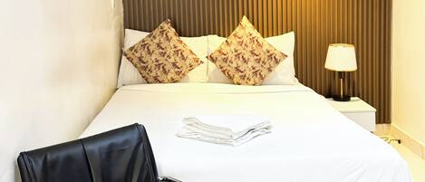 Una camera d'hotel con un letto, cuscini, una sedia, un comodino con una lampada e un quadro appeso al muro.
