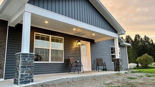 A modern house with a dark siding exterior, a stone pillar, and a porch with two chairs.