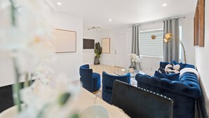 Een moderne woonkamer met een blauwe bank, een eettafel met stoelen en een groot raam met gordijnen.