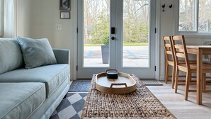 A living room with a grey sofa, a wooden dining table with chairs, and a large glass door leading outside. - Little Bell's Bungalow / Walkable to Saugatuck and Douglas / Updated (Saugatuck)