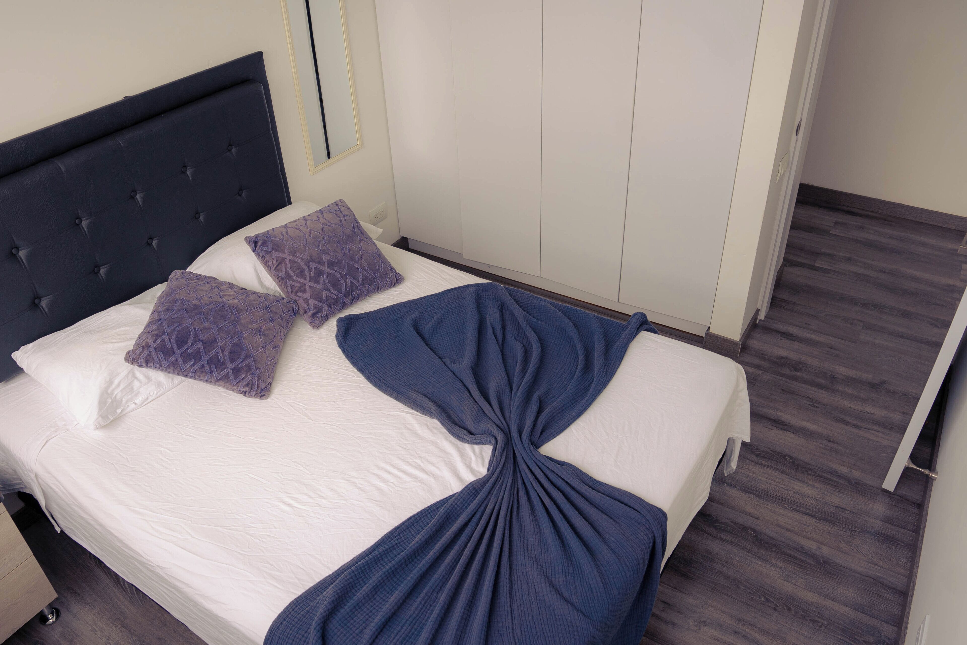 Ein ordentlich bezogenes Bett mit weißer Bettdecke, zwei violetten Kissen und einer dunkelblauen Decke, sowie ein dunkelblaues Kopfteil.
