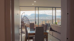 A modern living room with a glass dining table, two chairs, a sofa, and a hanging chair. A large window offers a city view with mountains in the distance.