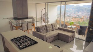 A modern kitchen with a gas stove, a glass dining table, and a sofa. There is a balcony with a view of a cityscape and mountains.