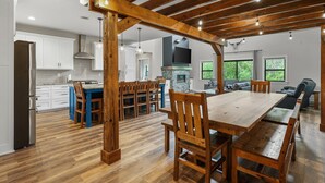 Uma cozinha ampla com vigas de madeira, um espaço de refeições com uma mesa grande e cadeiras, e vista para o exterior pelas janelas.