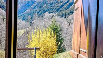A wooden barn with a metal railing, a yellow flowering tree, and a mountainous landscape with snow-capped peaks in the background.
