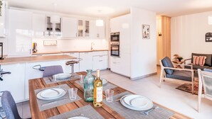 Una cocina moderna con armarios blancos, una mesa de comedor de madera para cuatro personas y un área de estar con sofá y sillones.