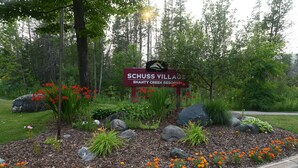 Skilt for Schuss Village Shanty Creek Resort omgivet af en anlagt have med sten og blomster.