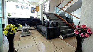 A modern living room with a sofa, coffee table, and two vases of flowers. A staircase with a wooden railing leads to an upper level. - Hotel A1 Plaza - Ahmedabad (Ahmedabad)