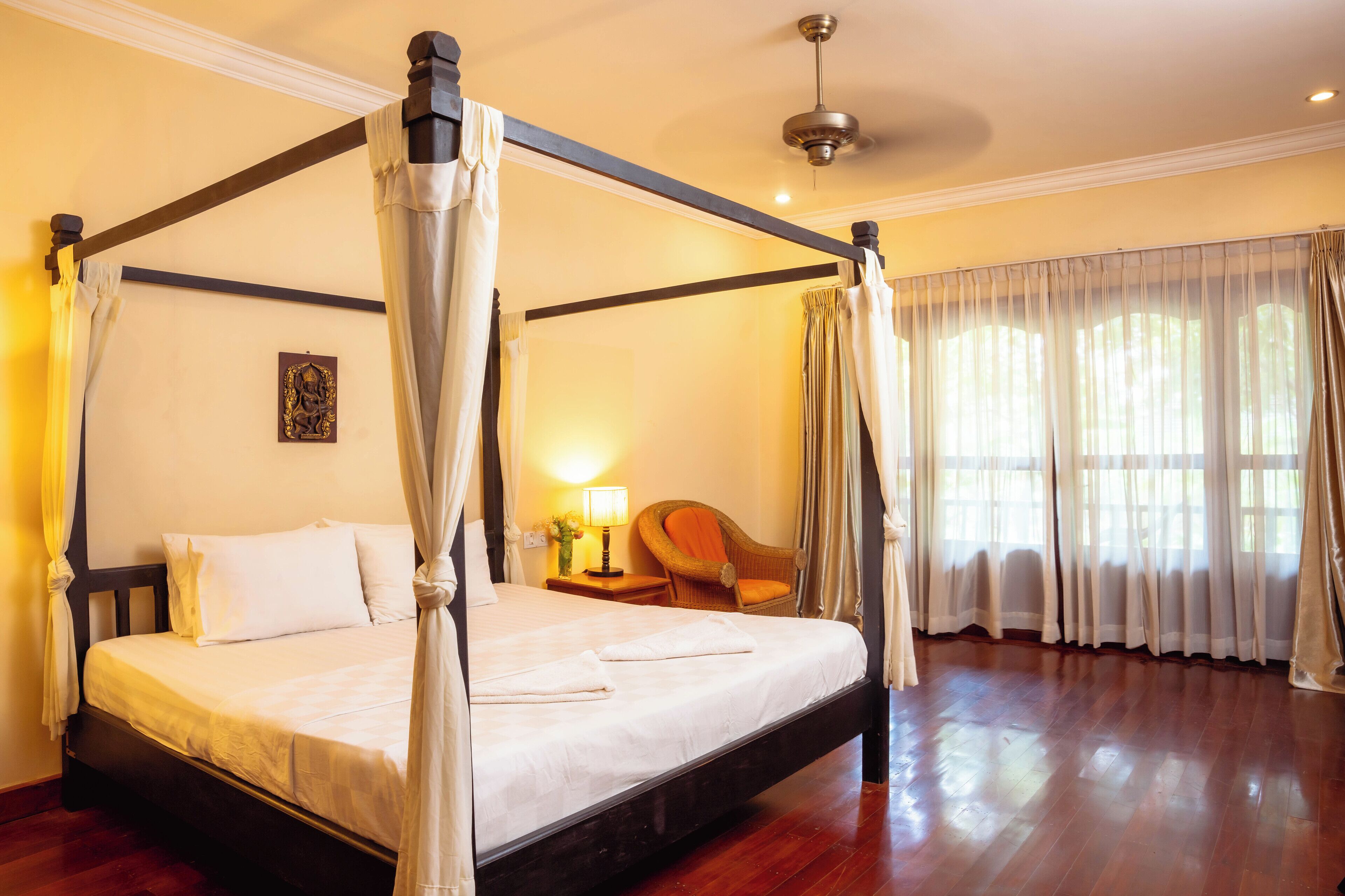 A four-poster canopy bed with white linens, a nightstand with a lamp, and a wicker chair.