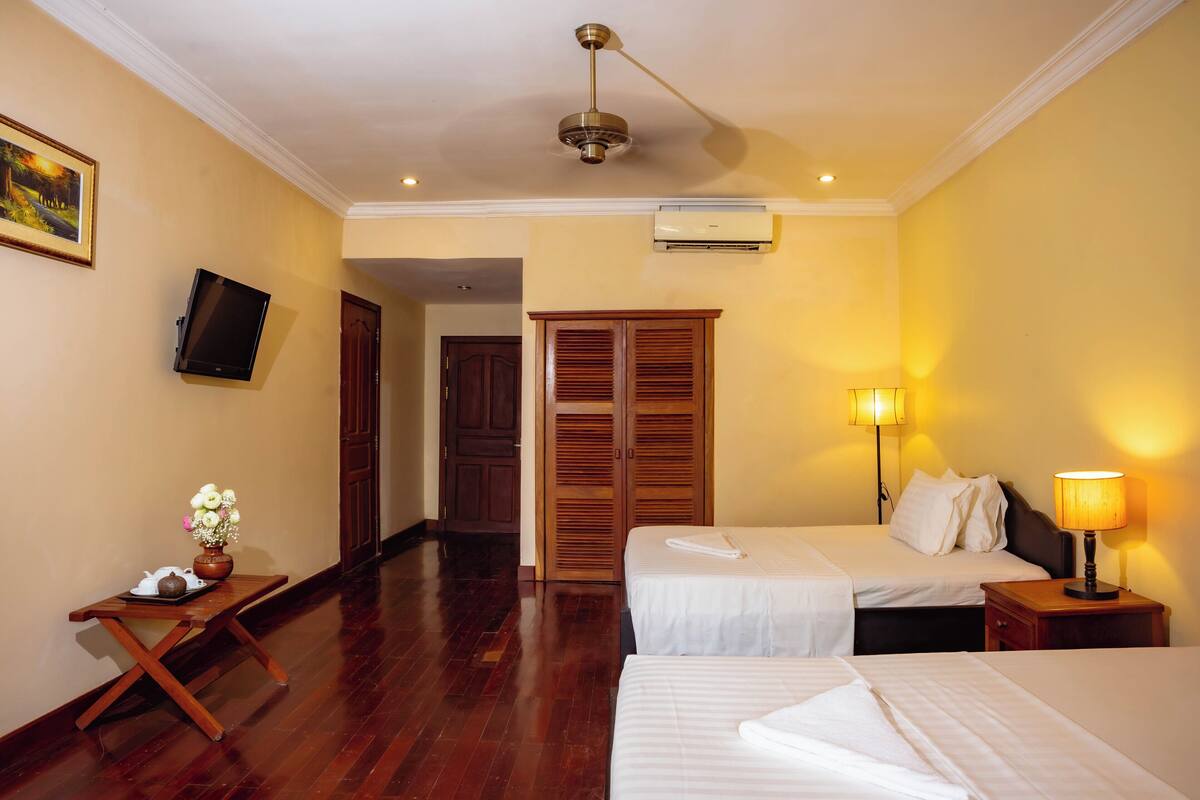 Quarto de hotel com duas camas, televisão fixada na parede, escrivaninha de madeira com um vaso de flores e ventoinha no teto.