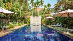 Una piscina con suelo de azulejos azules, rodeada de palmeras y sillones de descanso bajo sombrillas.