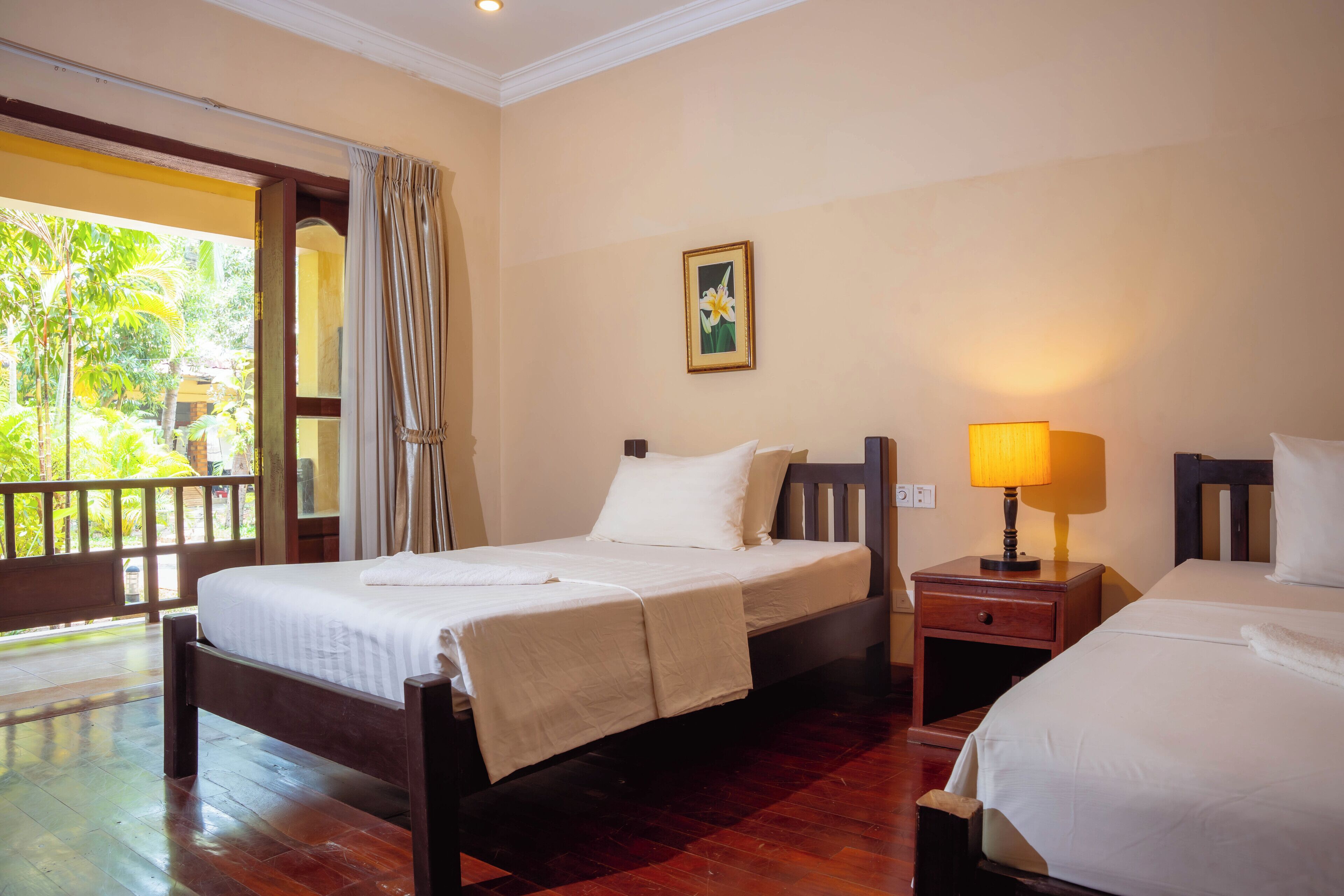 A hotel room with a wooden bed, a bedside table with a lamp, and a view of greenery outside.