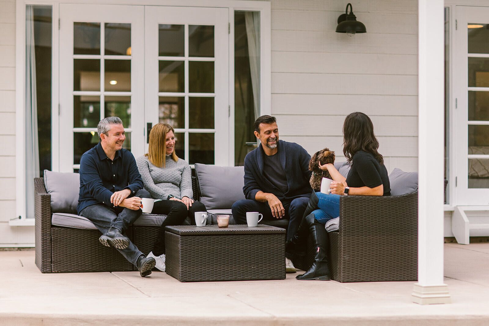 A group of people seated on a grey wicker sofa with a white cushion, in front of a white building with large windows and a black outdoor light fixture.
