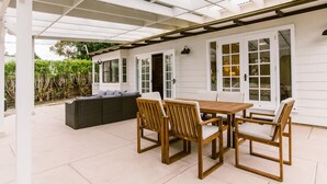 A covered patio with a wooden table and chairs, a dark sofa, and a white exterior wall with large windows.