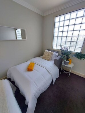 A bedroom with a bed, a bedside table with a plant, a window with blinds, and a mirror on the wall.