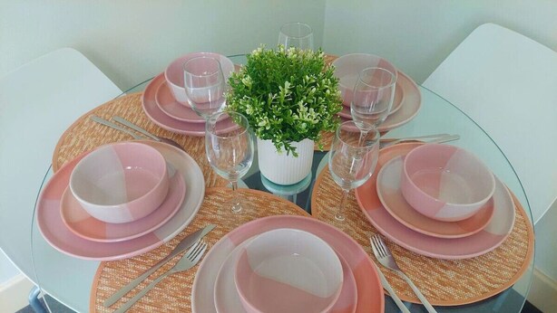 A round glass table with pink plates and clear glasses, surrounded by woven placemats and a small potted plant.