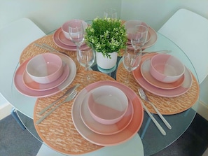 Una mesa de vidrio redonda con platos rosas y vasos transparentes, rodeada de individuales tejidos y una pequeña planta en maceta.