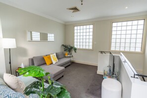 Una sala moderna con un sofá gris, una mesita auxiliar con una planta y ventanas amplias.