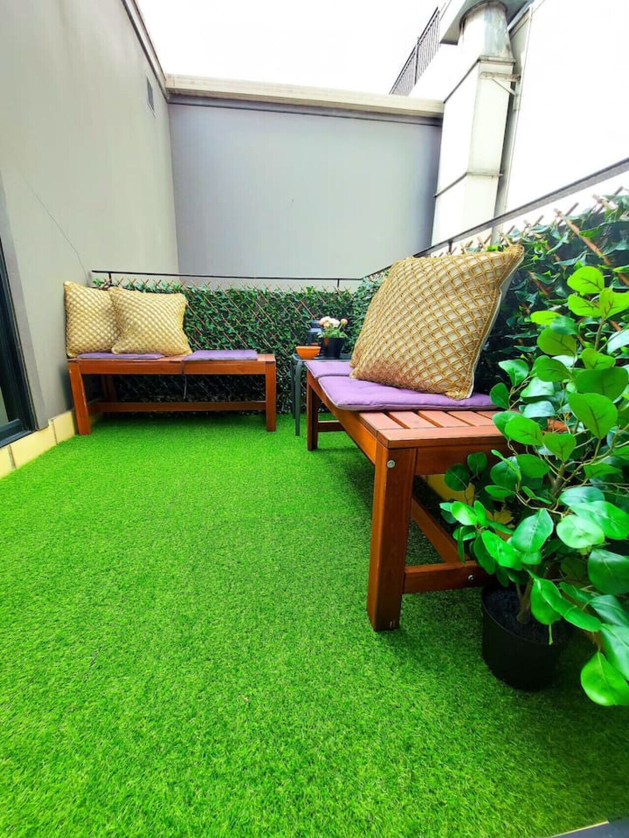 A small patio area with artificial green turf, two wooden lounge chairs with cushions, and a potted plant.