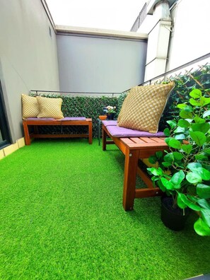 A small patio area with artificial green turf, two wooden lounge chairs with cushions, and a potted plant.
