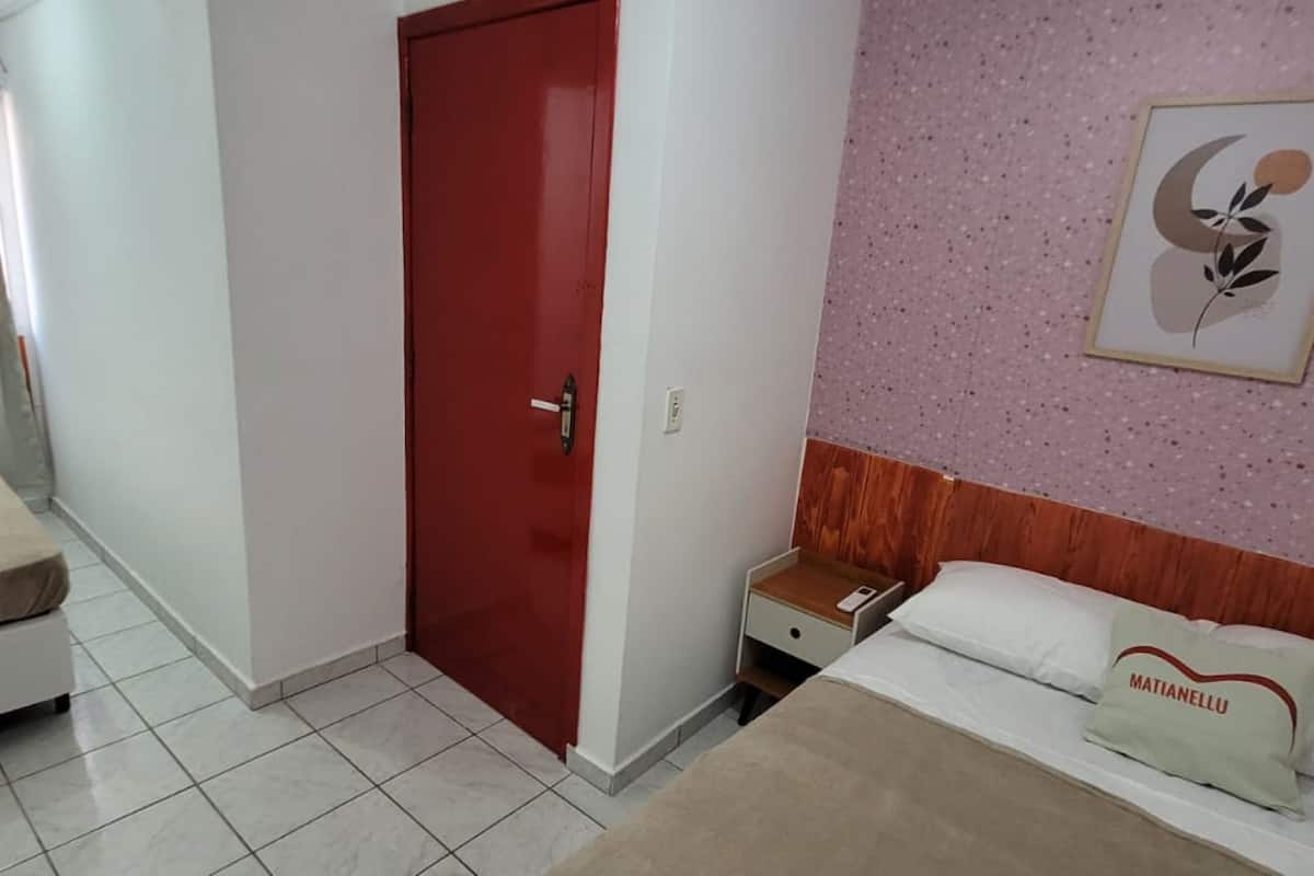 Una habitación de hotel con cama, mesita de noche, puerta roja y un cuadro en la pared.