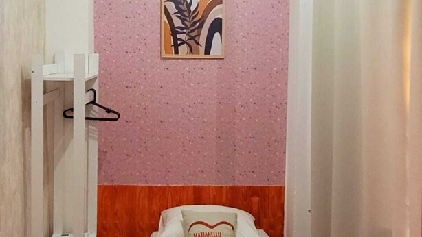 A small, single-bed room with a pink textured wall, a wooden headboard, a bed with a pillow labeled "MATIANELLO," a white shelf unit, and a shower curtain. - Matianellu SP (São Paulo)
