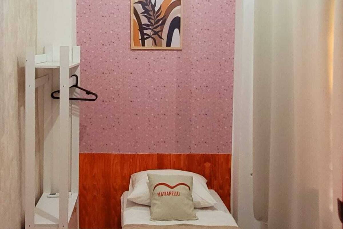 Una habitación pequeña con cama individual, pared de textura rosa, cabecero de madera, cama con un almohadón que dice "MATIANELLO", un estante blanco y una cortina de ducha.