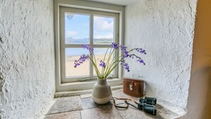 A window with a view of a beach, a vase with purple flowers, and a vintage camera on the windowsill. - Hen Dafarn -  an on the beach that sleeps 5 guests  in 3 bedrooms (Nefyn)