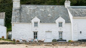 A white building with a grey tiled roof, two chimneys, and a stone wall in front. There are three wicker chairs and a small table outside. - Hen Dafarn -  an on the beach that sleeps 5 guests  in 3 bedrooms (Nefyn)