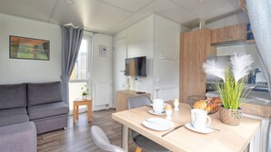 Una sala de estar moderna con un sofá gris, una mesa de comedor para dos personas y una zona de cocina con armarios de madera y un televisor.