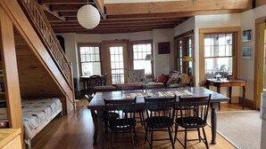 A spacious living room with a wooden staircase, a dining area with a long table and chairs, and large windows providing natural light. - Stunning cottage on Cayuga Lake, near wineries, waterfalls, Cornell University  (Interlaken)