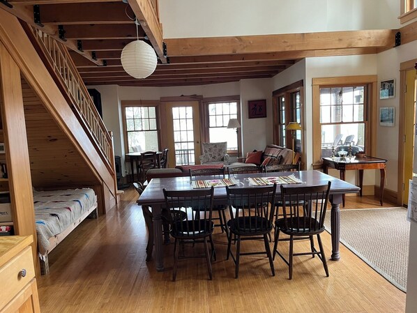 A spacious living room with a wooden staircase, a dining area with a long table and chairs, and large windows providing natural light.