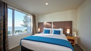 A hotel room with a large bed, two bedside tables, a view of the sea, and a window with curtains. - Kaikoura Luxury Apartments (Kaikoura)