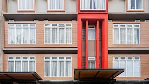 Un edificio de varios pisos con una estructura vertical roja en el centro, varias ventanas con cortinas blancas y un techo plano.