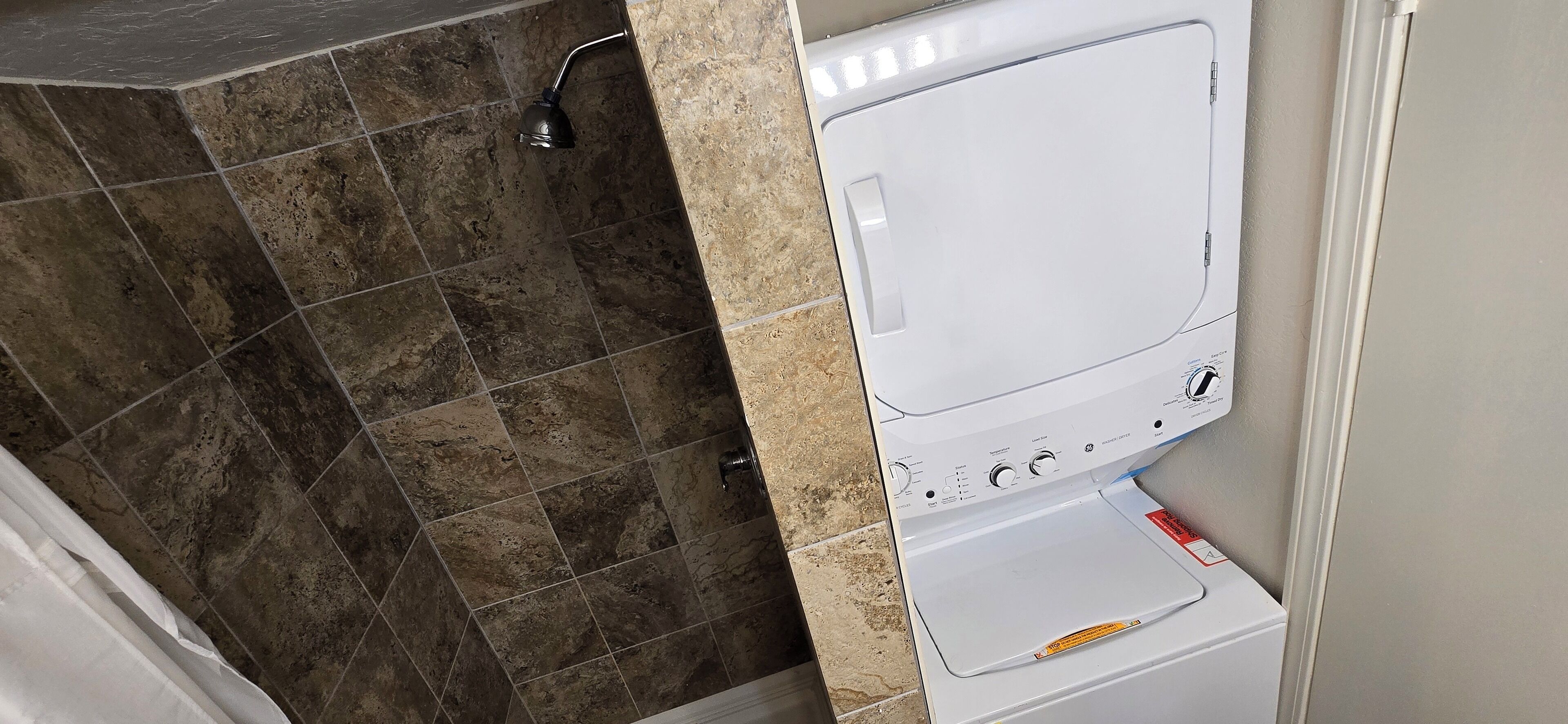 A bathroom with a white washing machine, a shower with a handheld showerhead, and a tiled floor.