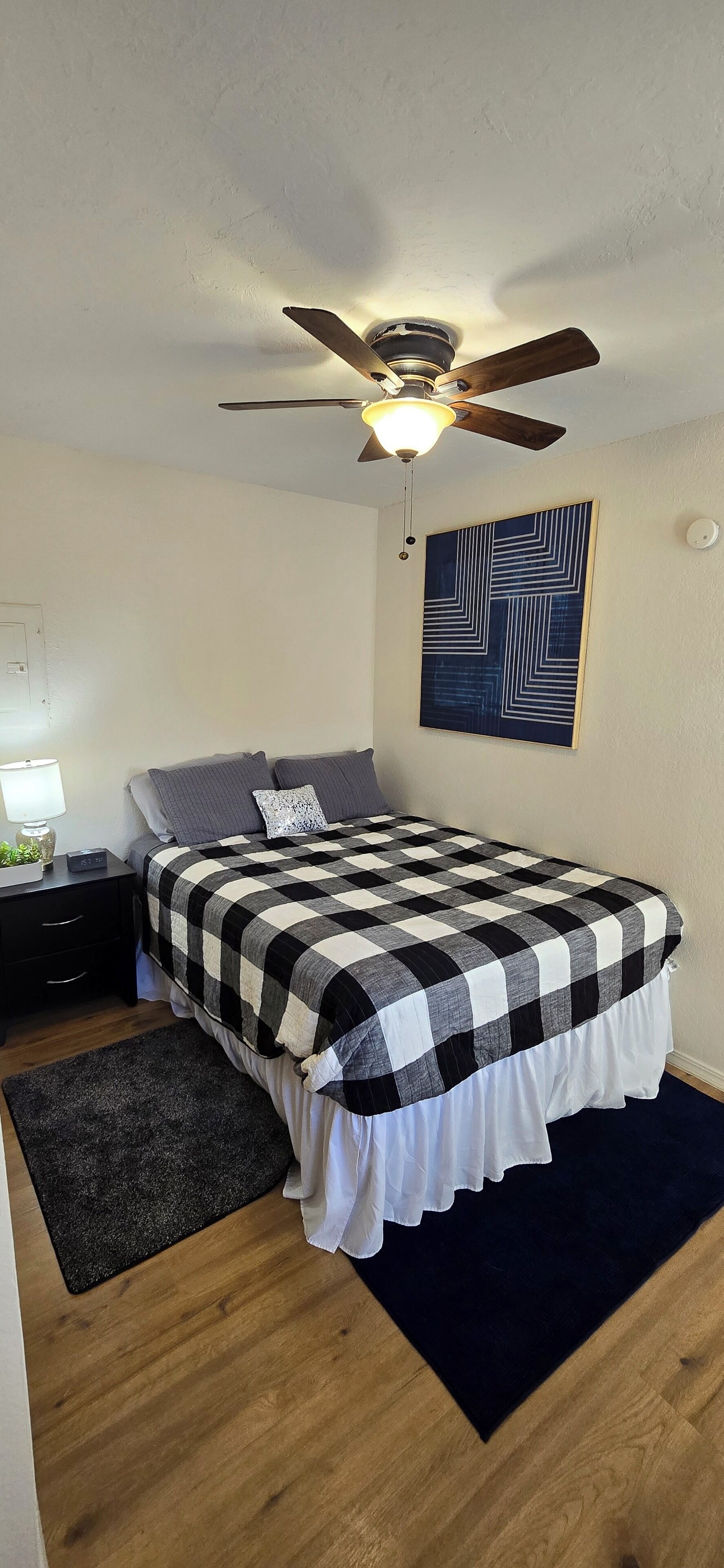 Ein Schlafzimmer mit Bett, Deckenventilator, Nachttisch, Lampe und Wandbild.