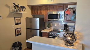 A compact kitchen with wooden cabinets, a stainless steel refrigerator, a microwave, a stove, and a small island with a decorative light. - Cozy Updated Cove of Heaven, 5 mins from Lake Hefner and mall. Centrally located (Oklahoma City)