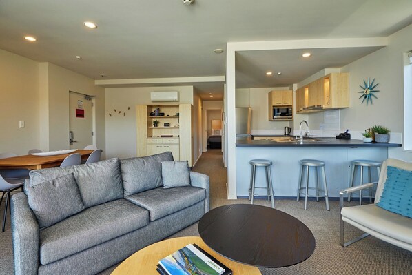 A modern living room with a grey sofa, a round coffee table, and a kitchen area with a bar stool. - Kaikoura Luxury Apartments (Kaikoura)