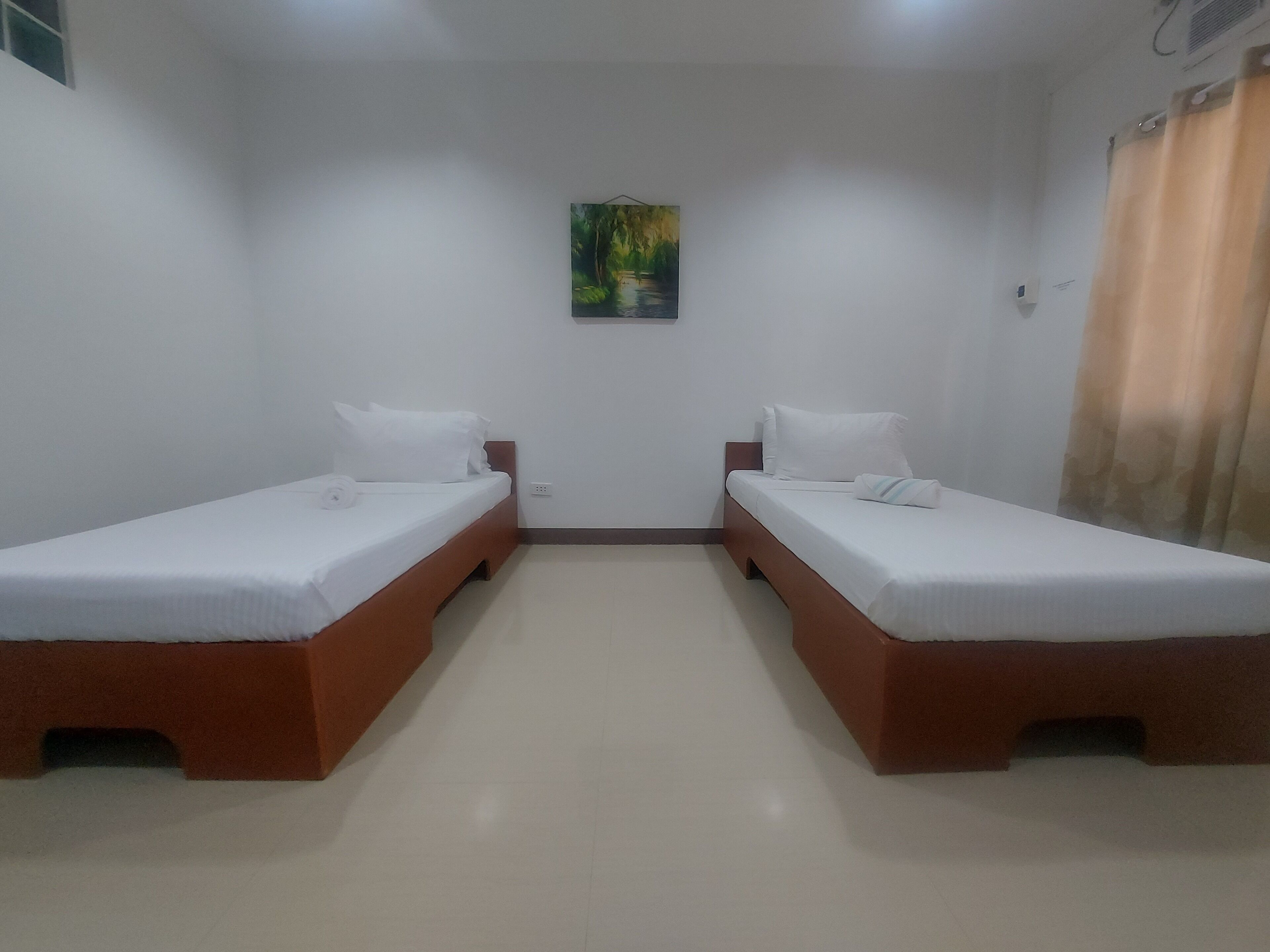 A room with two single beds, white bedding, wooden headboards, a small painting on the wall, and a window with curtains.