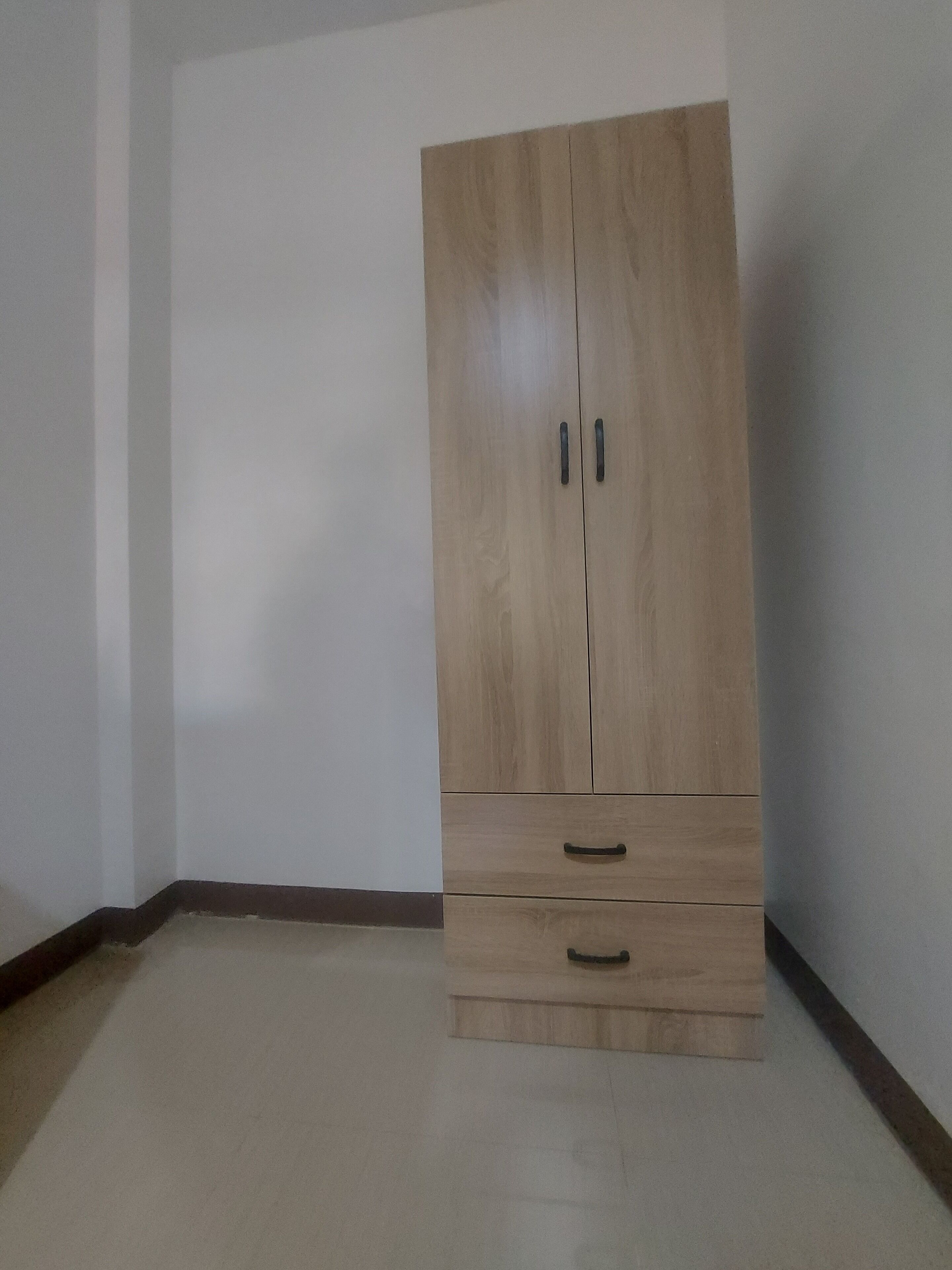 A wooden wardrobe with two drawers and two doors, positioned in a corner of a room with plain white walls and a tiled floor.