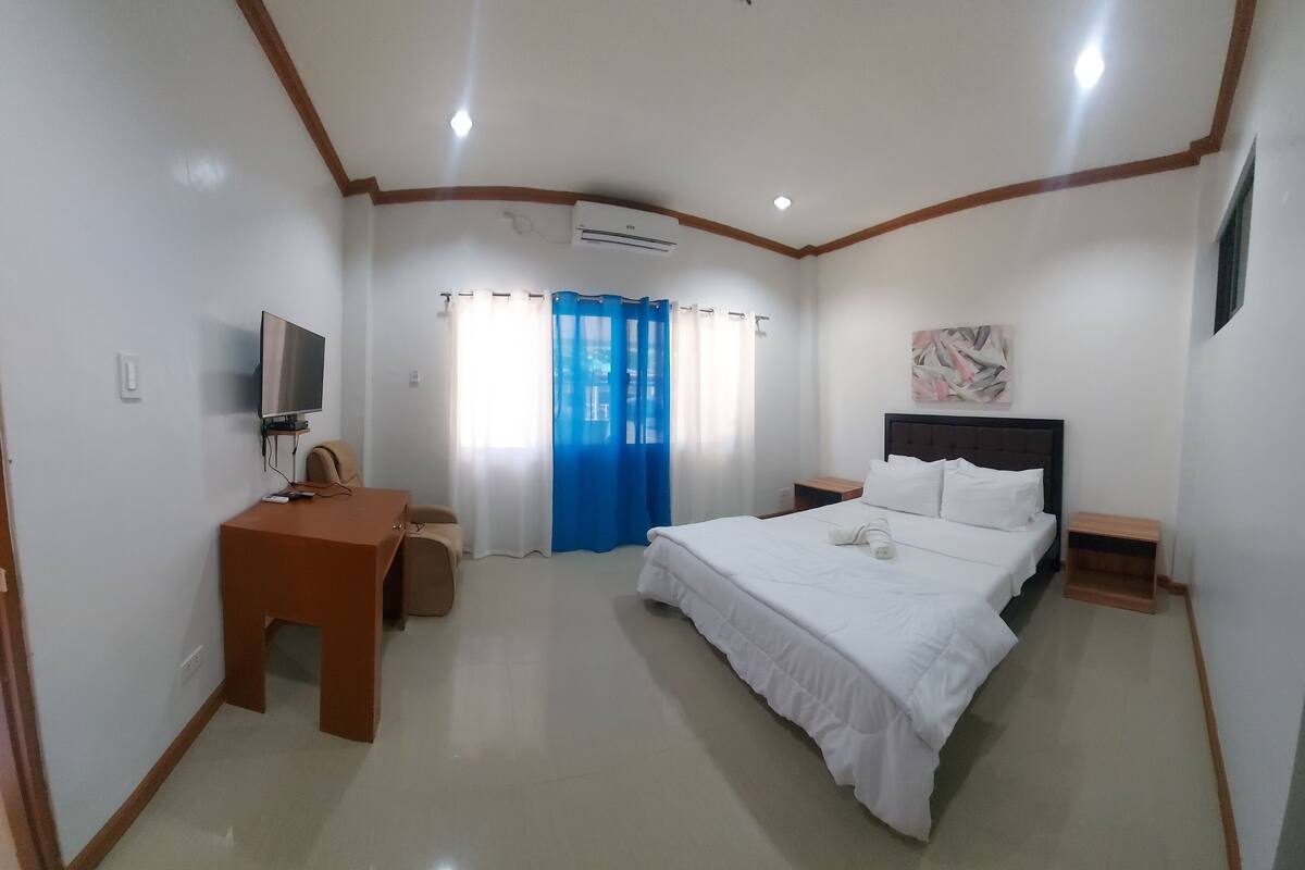 A hotel room with a bed, a desk, a chair, a TV, and an air conditioning unit.