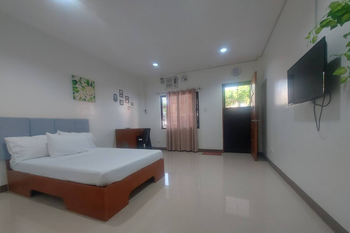 A bedroom with a bed, a desk, a chair, a TV, and a window with curtains.