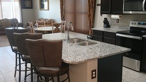 A modern kitchen with a central island, bar stools, a dining area with a sofa, and a ceiling fan. - Peaceful home in Estrella Mountains with beautiful lake for walking  (Goodyear)