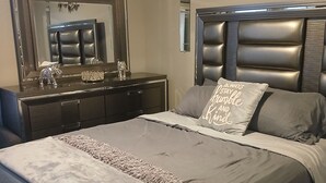 A bedroom with a large bed, a dresser, a mirror, and a decorative wall piece. - Peaceful home in Estrella Mountains with beautiful lake for walking  (Goodyear)