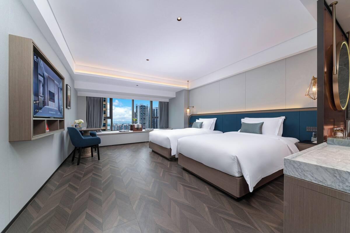 A modern hotel room with two beds, a desk, and a large window offering a city view.
