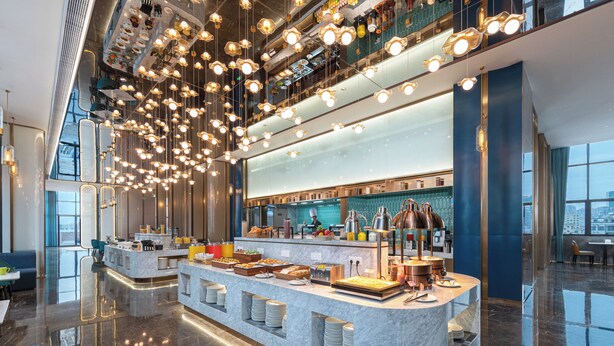Un moderno hall de hotel con una barra de desayuno central, techos altos y un suelo reflectante.