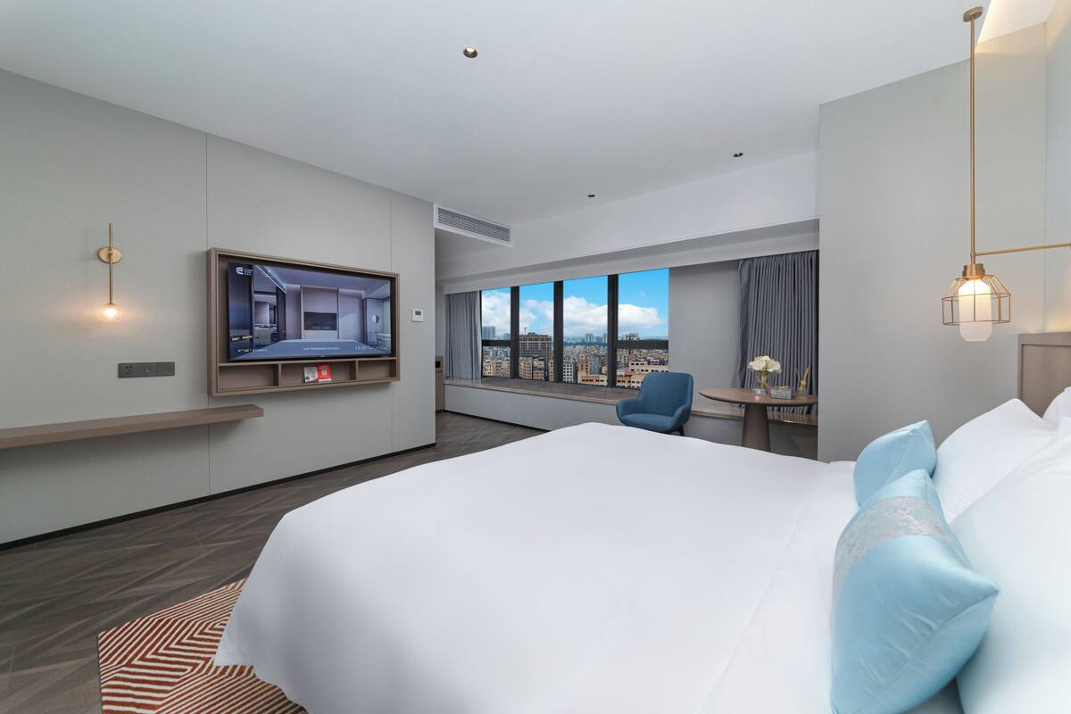 A modern hotel room with a large bed, a TV mounted on the wall, a city view through the window, and a small table with a chair.